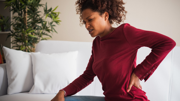 A woman in a red sweater sits on a white couch. Her hand rubs her lower back as she winces in pain.