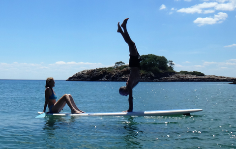 A man performs a handstand on a paddleboard in the ocean, while a woman sits and watches.