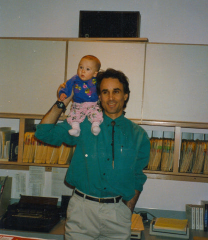 A man in stands in front of a wall of medical records with an infant girl on his shoulder.