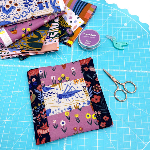A needle book on a cutting mat with some fabric and embroidery scissors.  There is a blue grasshopper on the front framed by a patchwork border of purple and black. 