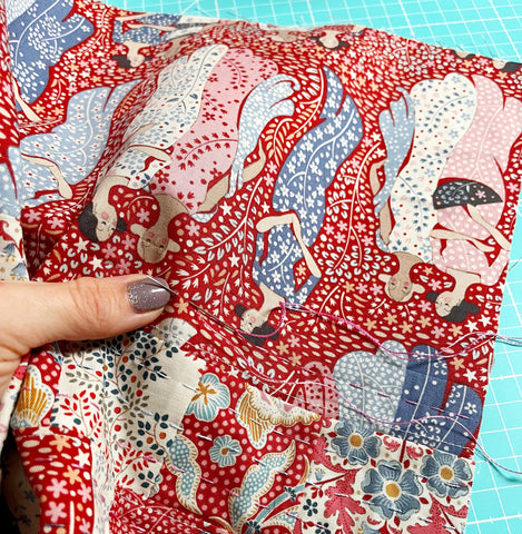 Hand stitching a running stitch along a patchwork piece that features a red background and ladies in long dresses. 