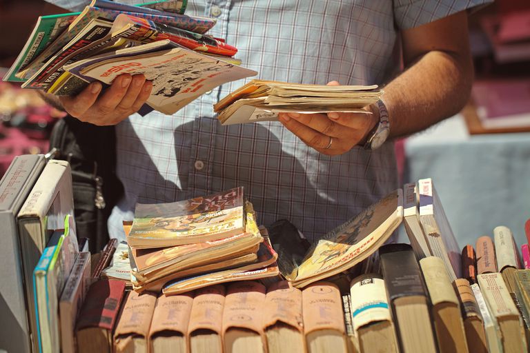 A man wearing a blue button up shirt holding comics while looking through a pile of books