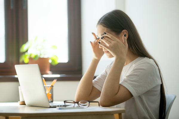 woman rubbing eyes sat at a table in front of a laptop