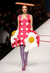 A female model wears a picnic blanket dress with fried egg decals, shiny purple leggings, and a baguette hat, 