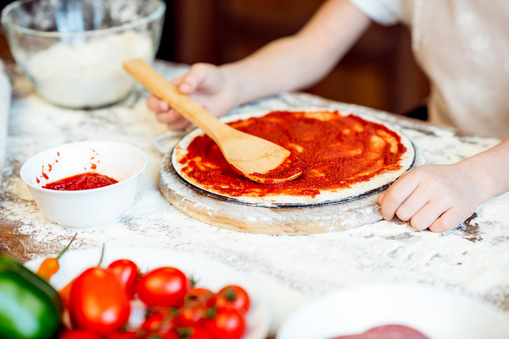 Partial view of girl putting tomato sauce on pizza dough