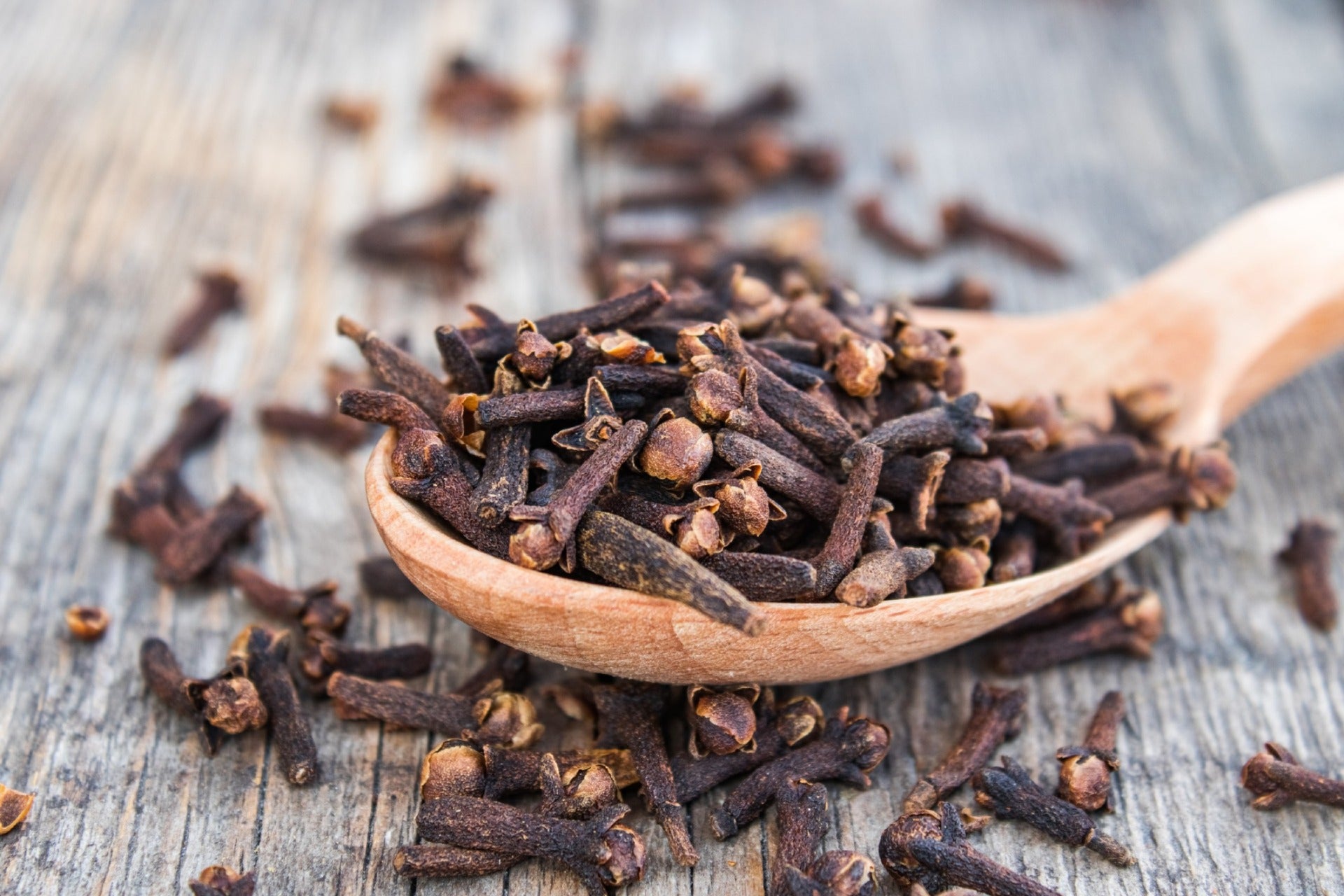 A cluster of dried cloves overflowing from a wooden spoon that lies on a wooden table.