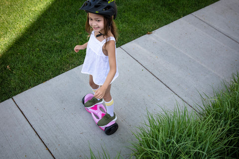 Young child riding a GOTRAX Pink SRX Mini Hoverboard for Kids with Built-In Carry Handles on a sidewalk