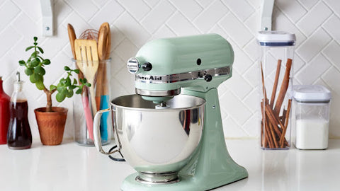 A green stand-alone mixer aid on top of a kitchen counter with some kitchen utensils and ingredients in containers in the background