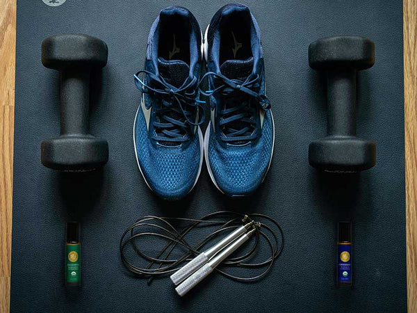 overhead photo, gym knolling: running shoes, barbells, jump rope, peppermint and eucalyptus essential oil roll-ons sitting on a yoga mat