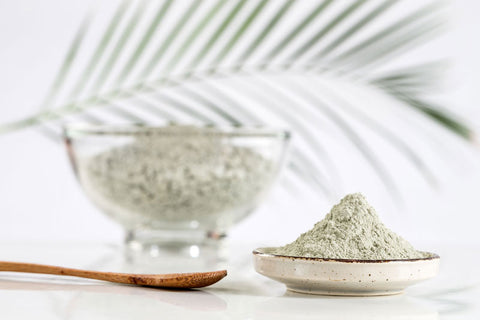 green clay powder in a bowl and small ceramic dish with bamboo spoon and palm frond in the background