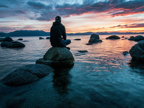 person sitting on a rock over water watching sunset