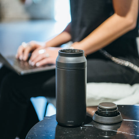 Kinto Travel Tumbler on desk while person works on laptop in coffee shop.