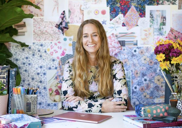 Alana owner of kukukachu design posing at her desk with all her patterns on the wall behind her