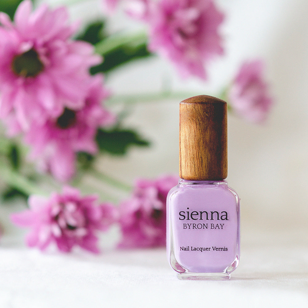 lilac nail polish bottle with timber cap and pink flowers in the background by sienna