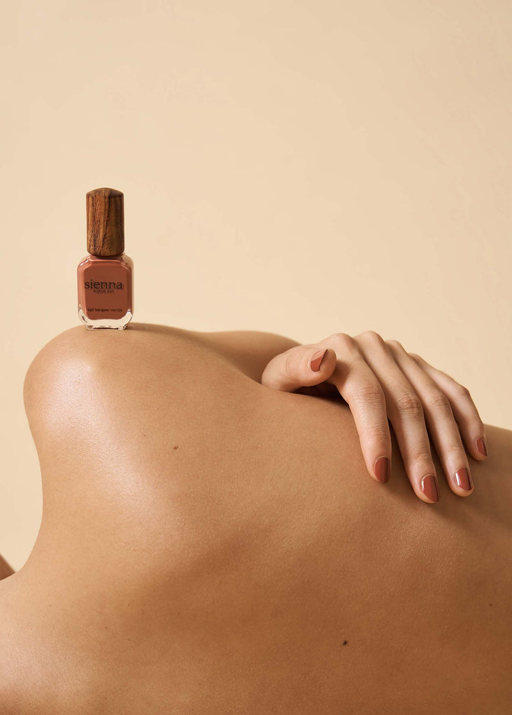 Woman lying down with tanned skin wearing Sienna nail polish with hand on back and nail polish bottle on shoulder.