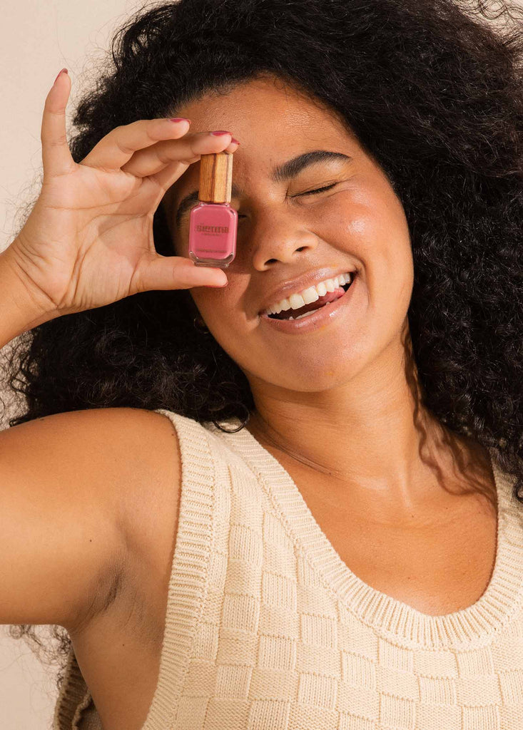 Medium brown skin woman wearing and holding Heartspace raspberry sorbet nail polish and holding to eye and smiling.