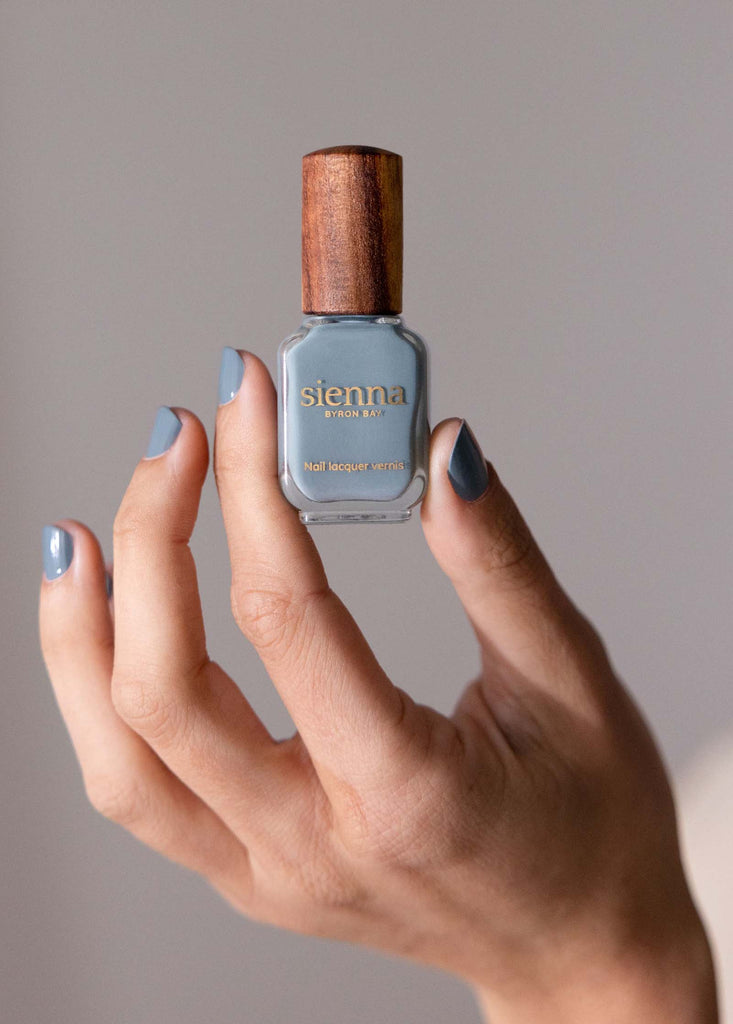 Fair skin hand wearing and holding juniper our gery-blue denim non-toxic nail polish by sienna with timebr lid. 