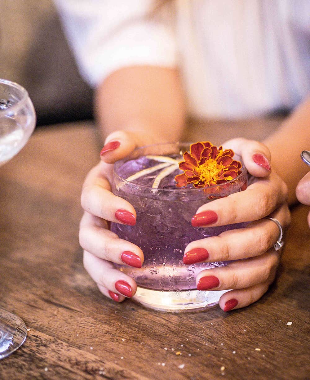 close view of a woman's hands holding a purple cocktail, wearing dusty rosebud red nail polish by sienna at Mez Club restaurant in Byron bay