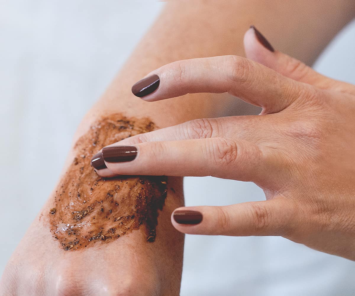 Close view of fair skin tone hands applying Cacao Brightener Vegan Face Mask, and wearing Deep brown umber nail polish by Sienna