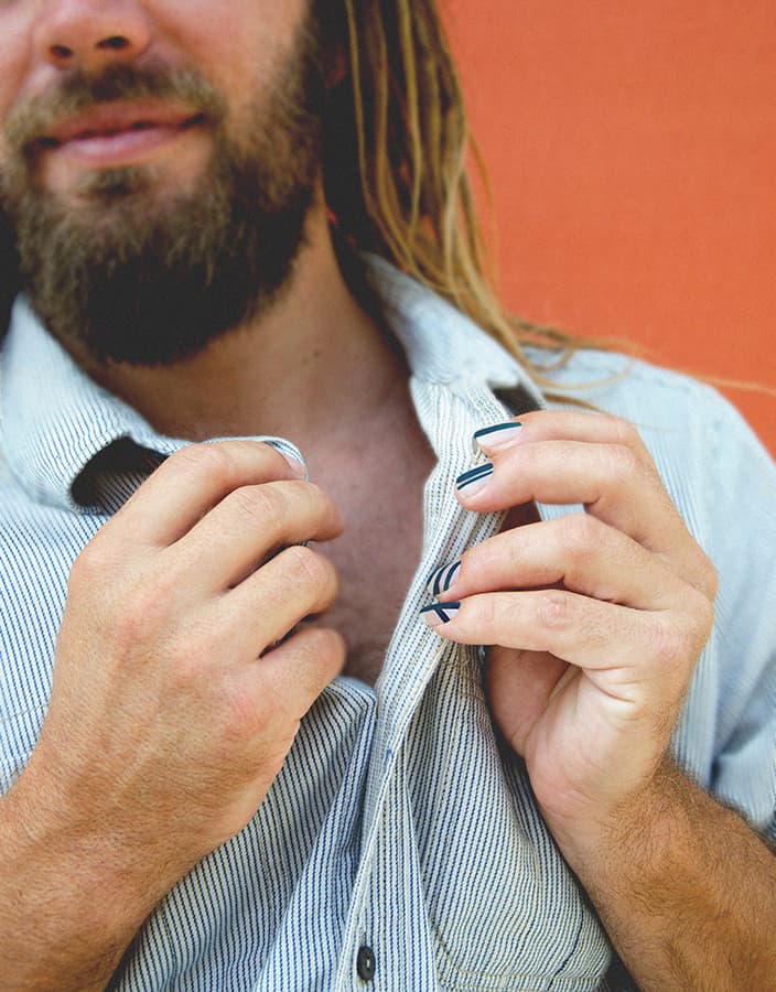 Close view of bearded man wearing teal blue nail polish in front of an orange wall holding his shirt collar