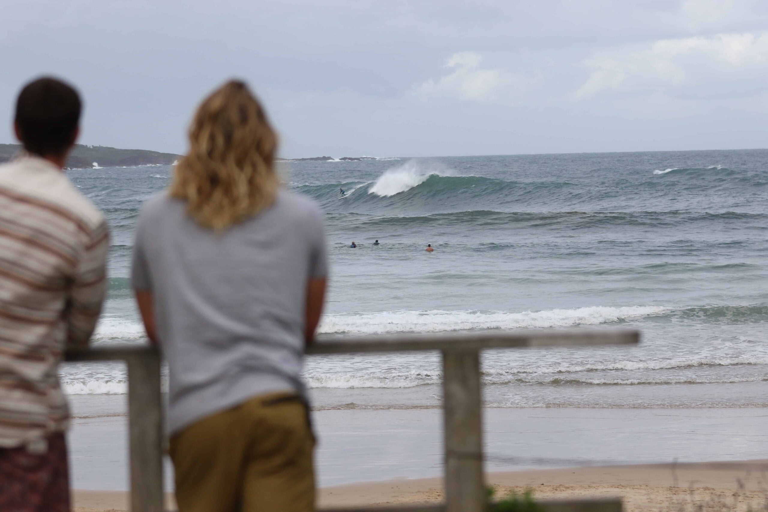 two men leaning on a wooden fence looking at the surfers and waves on a beach in australia