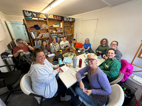 a big group of people holding knitting sitting around a table at Seattle Yarn, smiling at the camera