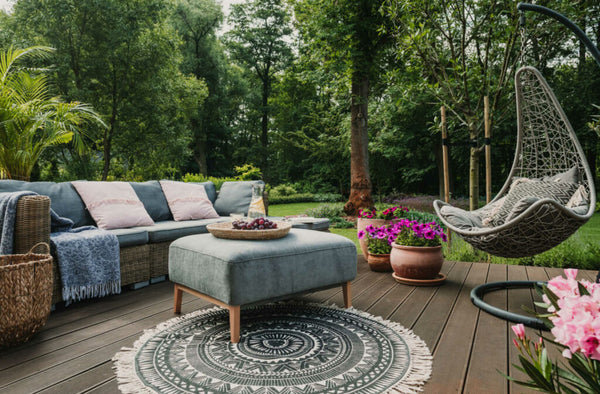 A garden patio decorated with a wicker sofa with cushions, a coffee table, and a round carpet with a vintage design.