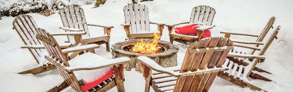 Patio chairs in winter piled with snow around a fire pit with a warm fire blazing