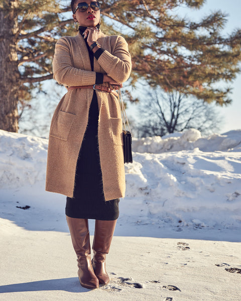 Fashion blogger Farotelle wearing a neutral outfit that consists of a black fitted turtleneck dress, a long camel cardigan with a belt, and tall brown leather boots. She has sunglasses on. There's snow in the background.