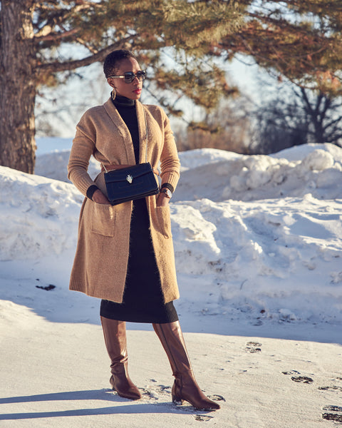Fashion blogger Farotelle wearing a neutral outfit that consists of a black fitted turtleneck dress, a long camel cardigan with a belt, and tall brown leather boots. She's also wearing a small black crossbody handbag. There's snow in the background.