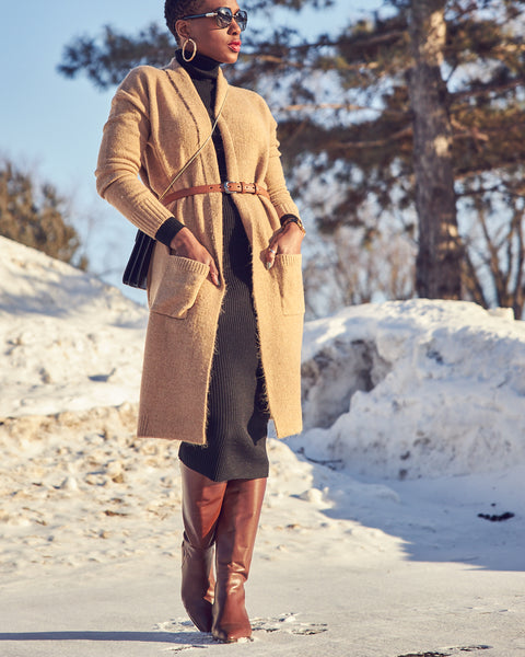 Fashion blogger Farotelle wearing a neutral outfit that consists of a black fitted turtleneck dress, a long camel cardigan with a belt, and tall brown leather boots. There's snow in the background.