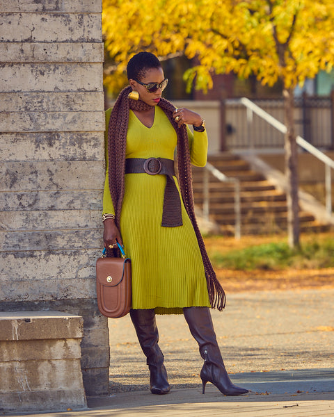 Fashion blogger Farotelle leaning against a stone wall and wearing a midi green sweater dress with a wide brown belt, a brown crochet sweater, and brown tall boots. She's holding a brown leather handbag and wearing sunglasses.