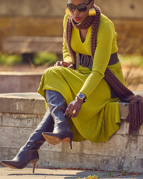 Fashion blogger Farotelle in a sitting position and wearing a midi green sweater dress with a wide brown belt, a brown crochet sweater, and brown tall boots. She is wearing sunglasses.