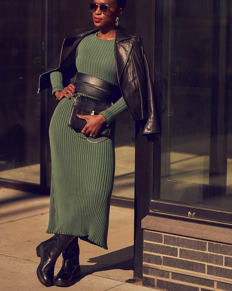 Fashion blogger Farotelle wearing a sage green sweater dress with a black wide waist belt, a black moto leather jacket, and black combat boots. She has sunglasses on and is wearing a black crossbody bag.