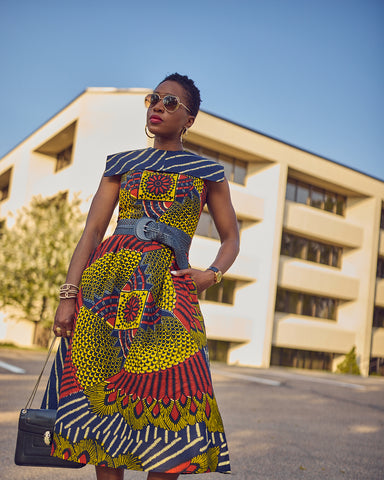 Style influencer Farotelle wearing a colorful Ankara midi dress. The dress has a distinctive blue and yellow print and a square collar. Farotelle is wearing the dress with a blue belt. There is a building with glass windows in the background.