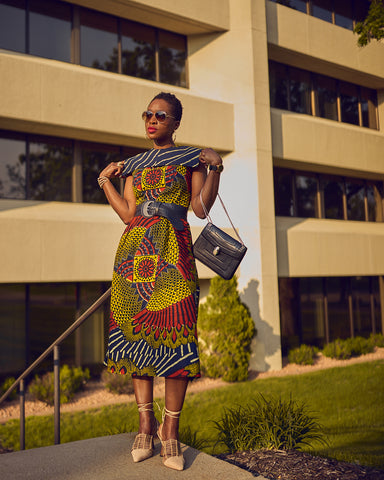 Style influencer Farotelle wearing a colorful Ankara midi Summer dress. The dress features a distinctive multi-colored print and has a square collar. Lace-up pumps and a blue belt complete the outfit. There is a building in the background.