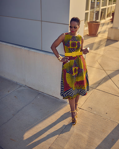 Style influencer Farotelle wearing a colorful printed midi dress. This is a sleeveless Ankara dress that is styled with a yellow belt and yellow strappy sandals. Farotelle has short black natural hair. She is standing against a wall.