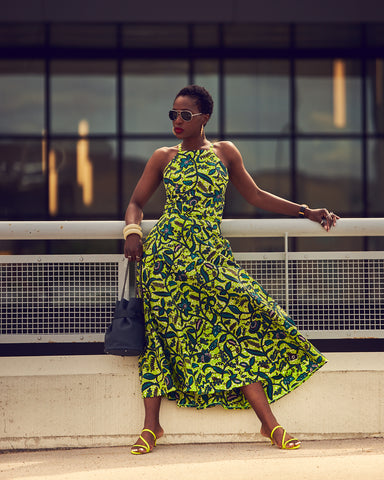 Style influencer Farotelle wearing a bright chartreuse Summer midi dress. The dress is sleeveless and has a floral pattern. She paired it with chartreuse strappy sandals and a blue bucket bag. She is leaning against guardrails and there are windows in the background.