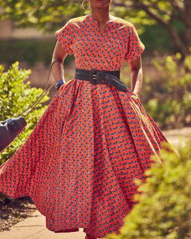 Style influencer Farotelle wearing a colorful orange maxi dress. This is an Ankara dress with a full skirt and a fitted top. Farotelle styled it with a contrasting wide blue belt. There's greenery in the background.