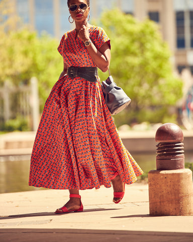 Style influencer Farotelle wearing a colorful orange maxi dress. This is an Ankara dress with a full skirt and a fitted top. Farotelle styled it with a contrasting wide blue belt and orange flat sandals. There's greenery in the background.