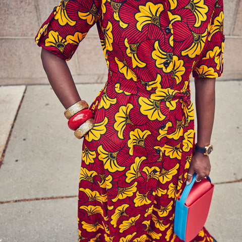 An outfit close-up picture showing style influencer Farotelle wearing a print dress. The dress has a red and yellow floral pattern. She accessorized it with colorful bangles and a color-blocked red handbag.