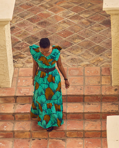 A top view of style influencer Farotelle wearing a colorful Summer maxi dress. The dress has flutter sleeves and a shell-patterned print in turquoise and brown. She completed the outfit with chunky black sandals by Vagabond and a black waist belt.