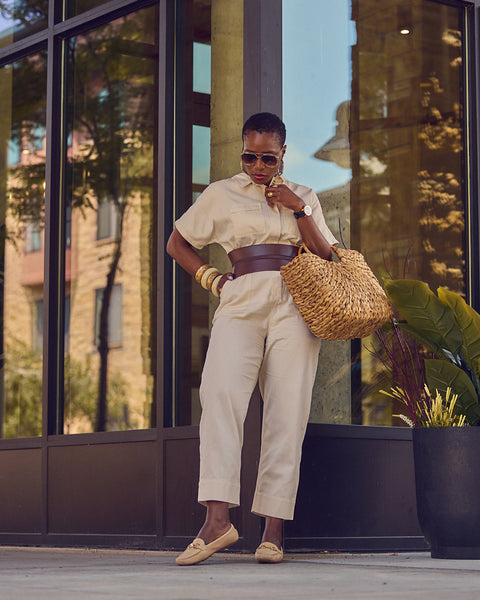 Influencer Farotelle posing against a glass background and wearing a beige utility jumpsuit with a brown waist belt, bangle bracelets, driver loafers, and a large straw bag.