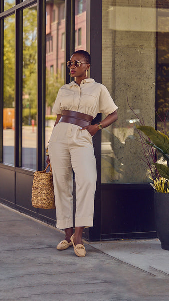 Influencer Farotelle posing against a glass background and wearing a beige casual jumpsuit with a brown waist belt, bangle bracelets, driver loafers, and a large straw bag.