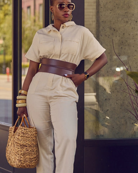 Influencer Farotelle standing against a wall and wearing a light beige jumpsuit with a wide brown waist belt, sunglasses, chunky bangle bracelets, and holding a large straw bag.