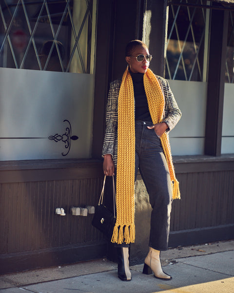 A woman wearing black ankle length jeans with a black turtleneck top and black and white plaid blazer. She has on color-blocked boots and a long yellow scarf.