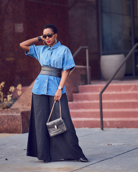 Style influencer Farotelle wearing black wide-leg linen pants with a belted denim shirt and black pumps. She is standing and is holding a black handbag. She has sunglasses on.