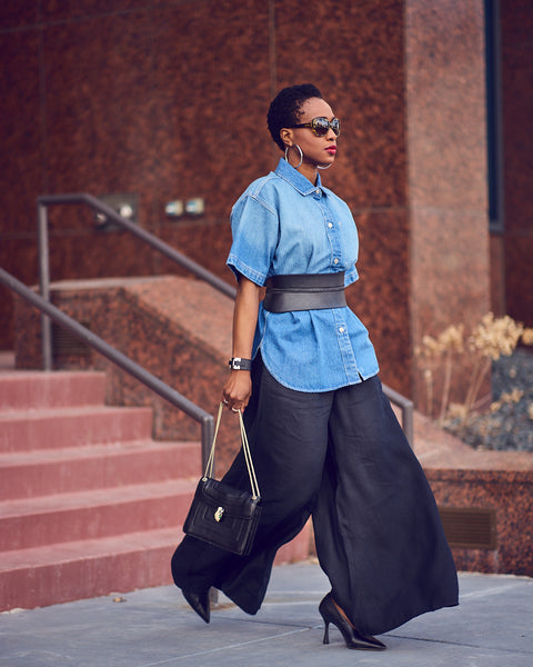 Style influencer Farotelle wearing black wide-leg linen pants with a belted denim shirt and black pumps. She is holding a black handbag and wearing sunglasses.