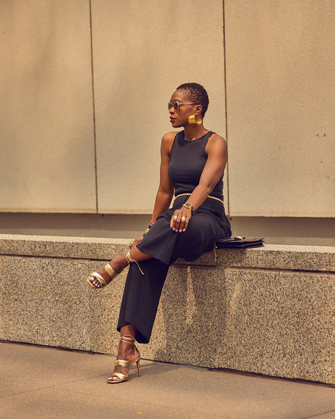 Fashion influencer Farotelle sitting on a wall and wearing an all-black outfit with gold heeled sandals and gold jewelry.