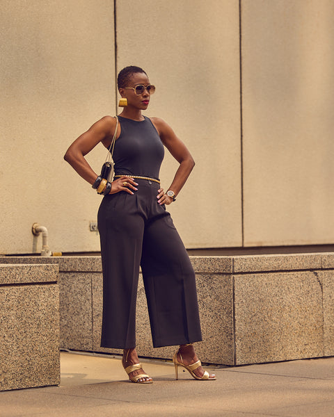 Fashion influencer Farotelle posing while wearing an all-black outfit with gold heeled sandals and gold jewelry.